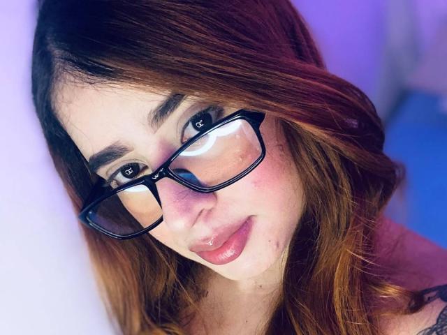 MartinaConnorr with glasses, pinkish skin, brown hair, slightly wavy, pink lips, subtle smile, looking at camera, blue eyes, soft light, purple-blue background, upper body, cute face.