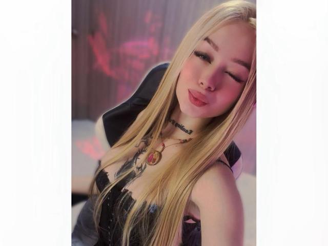 MiaValenttine with long blonde hair, pink eyeliner, and a black corset, showing off her tits and a tattoo on her chest, wearing a necklace with a pink gem.