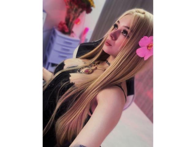 MiaValenttine with long blonde hair, black dress, pink flower in hair, tattoos on chest, looking at camera, slight smile, soft lighting, blurred background. Boobs visible, subtle cleavage.