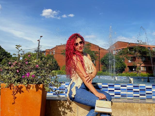 Antonela Riveiro, con cabello rizado rojo, gafas de sol y una blusa color piel, sentada en un borde de azulejos azules y blancos, sosteniendo un bolso beige, al lado de una maceta naranja con flores rosas..