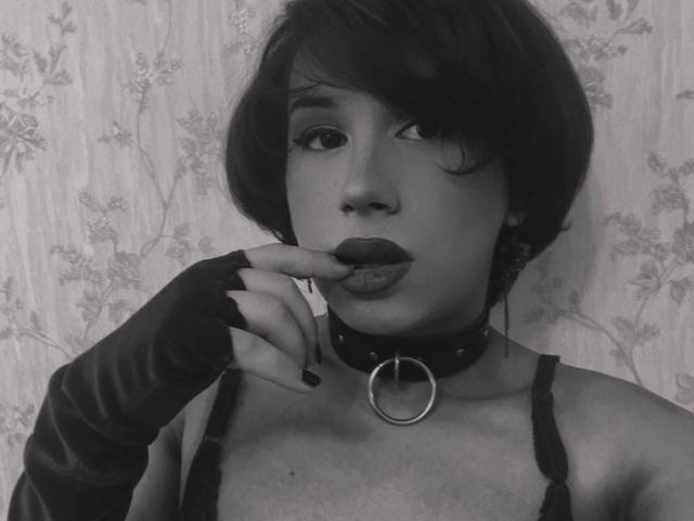 Fotografía en blanco y negro de FemCupid con cabello corto y oscuro, vistiendo un collar con un anillo, guantes sin dedos y una blusa negra, chupándose el dedo con los labios fruncidos..