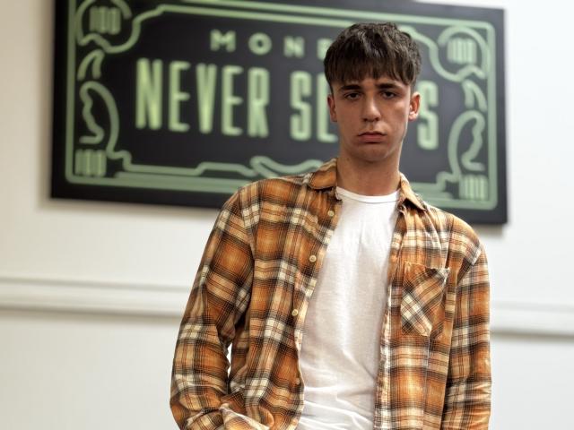 KellenWest with brown hair, wearing a yellow plaid shirt over a white tee, looking serious, hands in pockets, standing in front of a green sign.