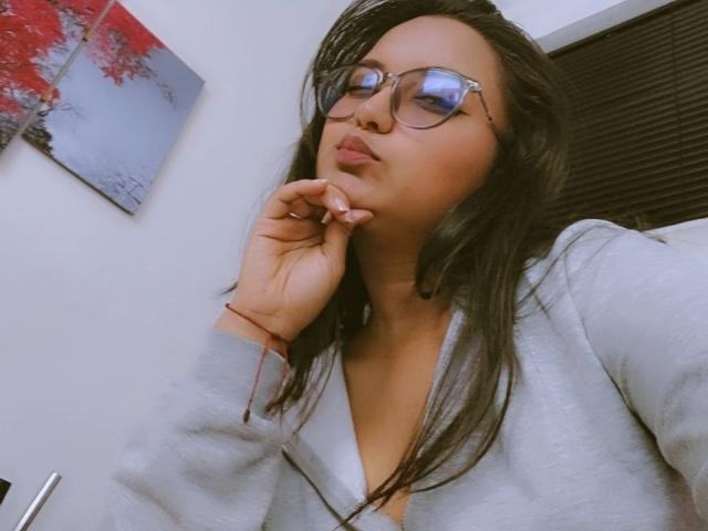 AzulMoon with pouty lips, wearing glasses, grey hoodie, and black hair, touching nose, showing cute, sexy face, with soft brown skin, in a casual selfie.