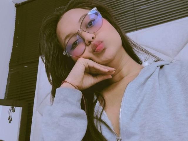 AzulMoon with puffy pink-tinted glasses, long dark hair, and a grey zip-up hoodie, puckering lips, resting chin on hand, showing soft, cute, and slightly nerdy vibe.