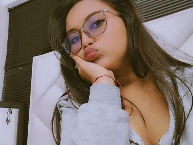 AzulMoon with puffy lips, wearing glasses, grey hoodie, hand on cheek, long brown hair, purple eyeshadow, cute face, medium tits, red bracelet, white headboard, brown blinds.