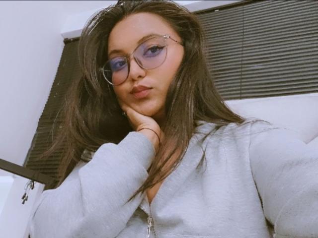 AzulMoon with long brown hair, wearing glasses and a gray hoodie, pouting with hand on chin, looking at camera. She has blue eyeshadow and a cute, serious expression.