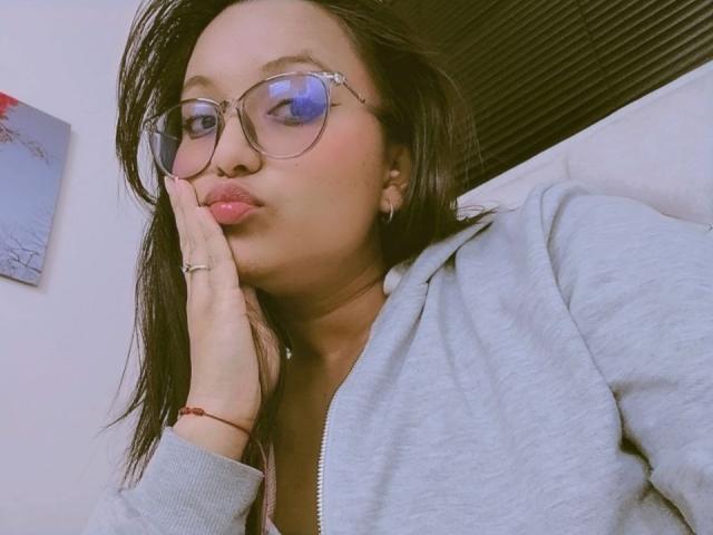 AzulMoon with pouty lips, wearing glasses, grey hoodie, and red bracelet, has black hair, hand on cheek, reflecting blue light in photo. Her tits are partially visible.