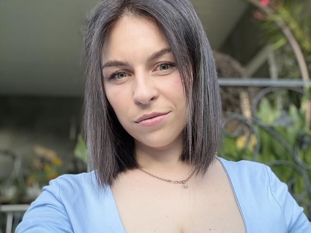EmmaNoa, con cabello negro de longitud media, ojos azules, piel clara y una sonrisa sutil, lleva una blusa azul claro y un collar, tiene senos pequeños y un rostro lindo..