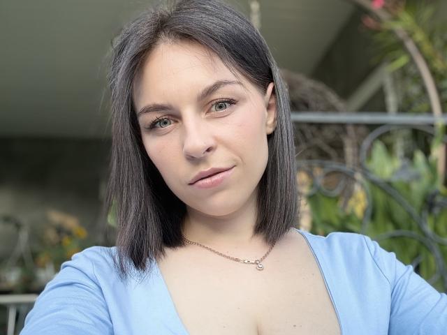 EmmaNoa hat eine blasse Haut, grüne Augen und dunkelbraunes, schulterlanges Haar. Sie trägt ein hellblaues Oberteil mit tiefem Ausschnitt, das etwas Dekolleté zeigt, und eine kleine Halskette..