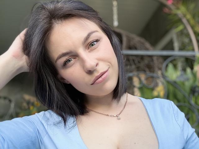 EmmaNoa with short black hair, light skin, and green eyes, wearing a light blue top, smiling, hand in hair, necklace, soft makeup, and pinkish lips.