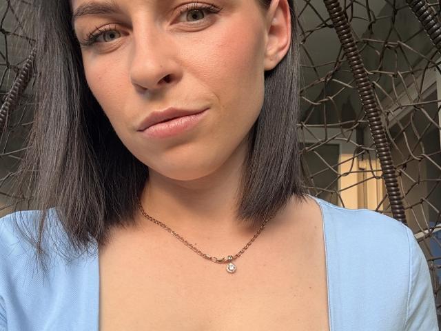 EmmaNoa mit glattem, schwarzem Haar, heller Haut und grünen Augen, trägt ein blaues Oberteil und eine zarte Halskette, die dezent das Dekolleté zeigt, und sie hat ein kleines Lächeln..