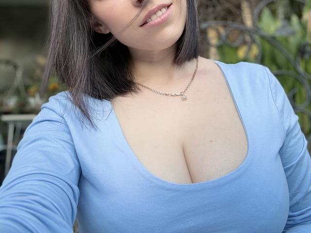 EmmaNoa, con un seno di media grandezza, indossa una maglietta blu, sorride, porta una collana d'argento, ha capelli castano scuro che le arrivano alle spalle e sta scattando un selfie, mostrando la parte superiore del suo corpo..