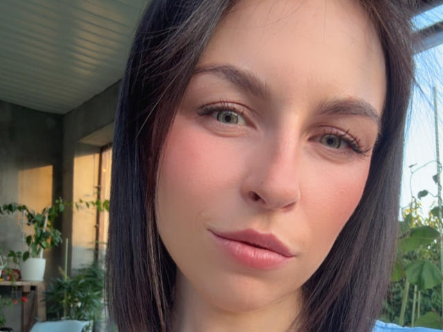 El rostro de EmmaNoa, con piel pálida, ojos verdes, cabello lacio de color marrón oscuro, labios rosados y un maquillaje sutil. Su nariz es pequeña, y está mostrando una ligera sonrisa, con una expresión de confianza..