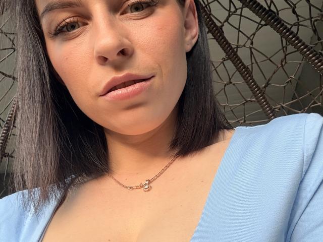 O rosto de EmmaNoa, com pele marrom média, cabelo preto liso e uma blusa azul clara, usando um colar delicado com uma pequena pérola, mostrando uma maquiagem discreta e um leve sorriso..