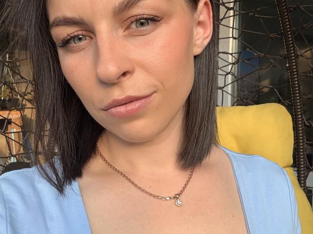 EmmaNoa, com pele clara, olhos verdes e cabelo preto curto, vestindo uma blusa azul e um colar marrom, sorrindo levemente. Seios não visíveis. Almofada amarela ao fundo..