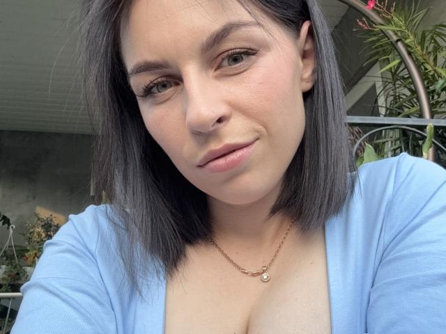 EmmaNoa, con un corte de pelo bob gris, piel clara, una blusa azul, un collar de oro pequeño, una sonrisa sutil, escote visible y senos pequeños, en un selfie con luz natural..