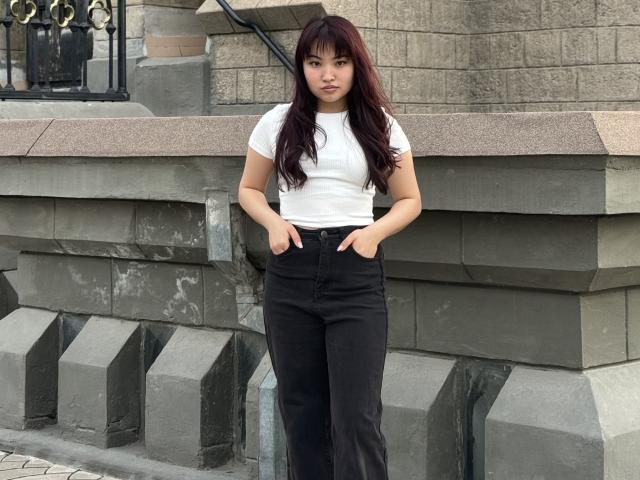 Leora Hardeman, met haar handen in zwarte broek, een wit T-shirt, lang donkerrood haar, staand tegen een stenen muur, kijkend naar de camera, met een lichte glimlach, middelgrote borsten, schattig, zelfverzekerd, casual..