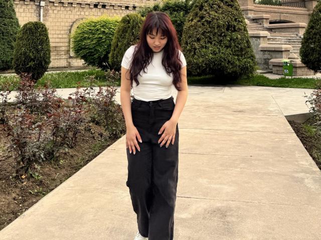 LeoraHardeman stands on a concrete path with hands on her black, loose-fitting pants, wearing a white, short-sleeved shirt, and white sneakers, with long, dark red, wavy hair.
