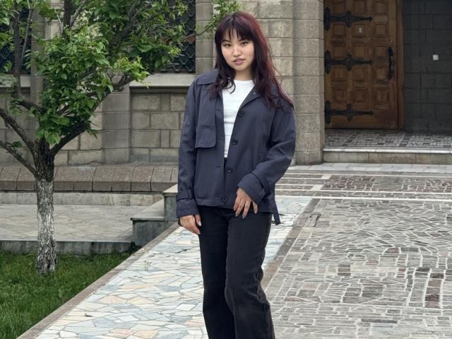 LeoraHardeman stands with dark red hair, wearing a black jacket over a white top, and black pants, in front of a stone building with a wooden door.