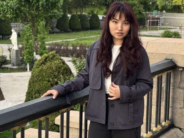 LeoraHardeman with long dark hair, wearing a black jacket and white shirt, standing by a black railing, looking at the camera with a neutral expression.