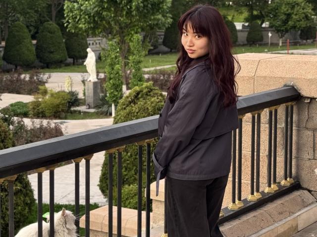 LeoraHardeman with long dark red hair, black jacket, and black pants, standing on a stone staircase, looking over her shoulder, hands in pockets, black railing with gold accents.