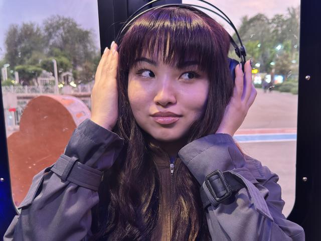 LeoraHardeman with brown hair, wearing headphones, gray jacket, and belt, hands on ears, smiling slightly, inside bus, evening light, trees and street visible outside window.
