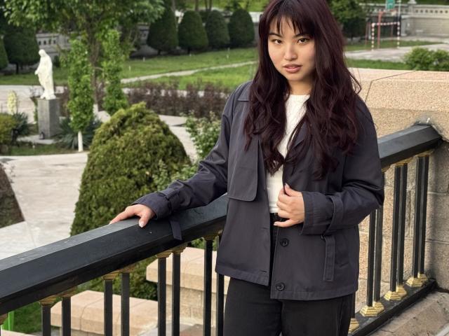 LeoraHardeman with long dark brown hair, wearing a black jacket and white shirt, standing against a black metal railing, looking at the camera with a slight smile.