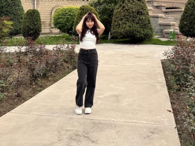 LeoraHardeman with hands in her black hair, wearing a white tank top and black pants, standing on a concrete path, white sneakers, small ass, medium tits, and a cute face.