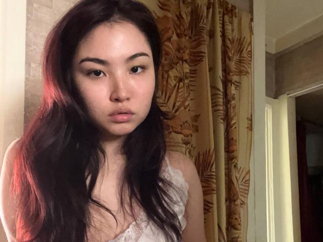 LeoraHardeman with long dark hair, wearing a white lace top, has a pouty face, and is looking directly at the camera. Her tits are small and she's indoors.