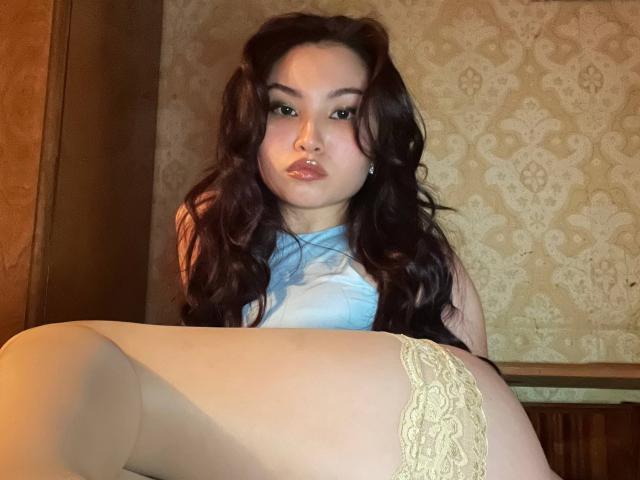 LeoraHardeman with long dark wavy hair, light blue top, and yellow lace panties, showing her white thigh and buttocks, looking at the camera with a slightly pouty expression.