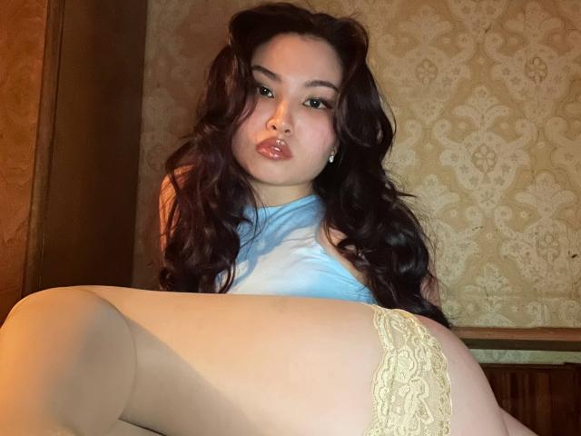 LeoraHardeman with long dark hair, wearing a blue top and yellow lace panties, has her ass in the foreground, pouting with full lips, looking at the camera.