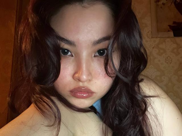LeoraHardeman with wavy brown hair, pouty pink lips, and dark eyebrows, staring into the camera, showing her pale skin and subtle freckles, wearing a blue top.