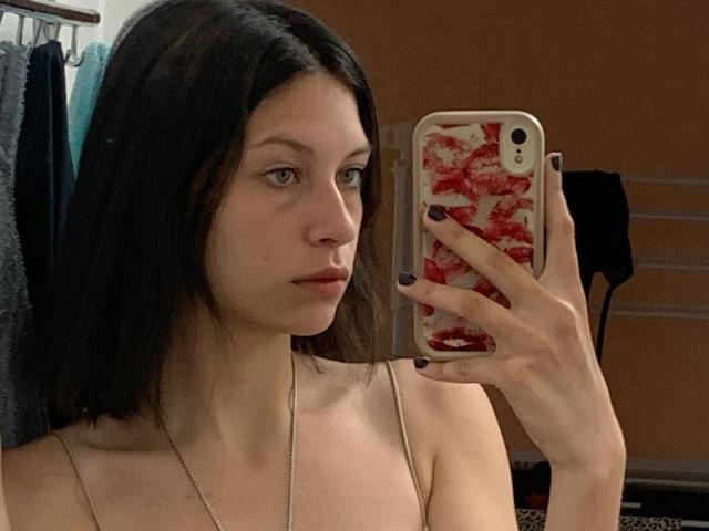 DavinaCegla with black hair, pale skin, and small tits, holding a phone with a red floral case, taking a mirror selfie, wearing a thin-strapped top, and black nail polish.