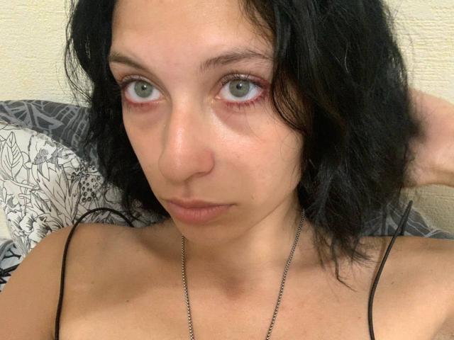 DavinaCegla tem pele clara, olhos verdes, cabelo preto ondulado e uma expressão neutra. Ela está vestindo uma blusa preta com alças finas e um colar de prata. Seus seios estão visíveis..