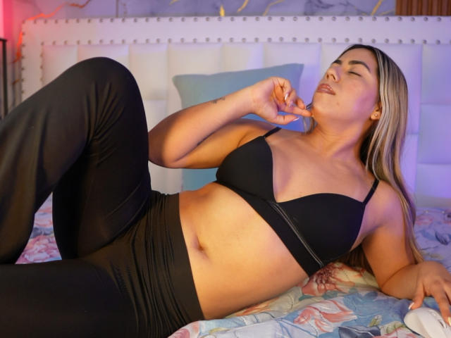 QuickHookup with light brown skin and blonde hair, lying on a bed in black sports bra and pants, fingers near mouth, eyes closed, showing her tits and slightly visible pussy.