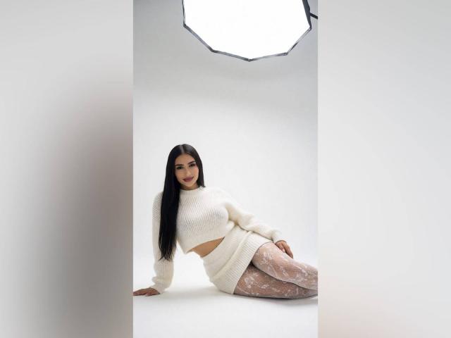 MiilaWx has long black hair, white crop sweater, and sheer patterned tights, sitting with her ass slightly raised, smiling at the camera in a studio with a white background.