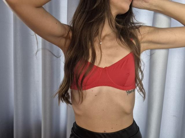 TenerliRize with long brown hair, arms raised, wearing a red bra, exposing slim, toned abs, small tattoo on right side, and black pants, standing in front of a curtain.