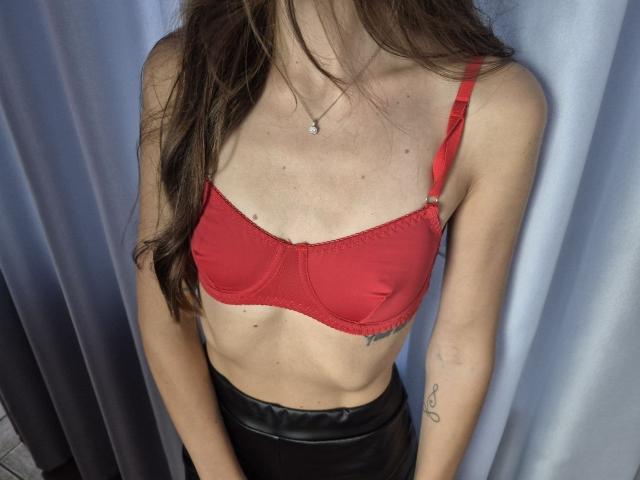 TenerliRize, avec un soutien-gorge rouge, montrant ses seins, de longs cheveux bruns, mince, ventre plat, pantalon en cuir noir, petit tatouage sur le bras droit, debout devant un rideau gris..
