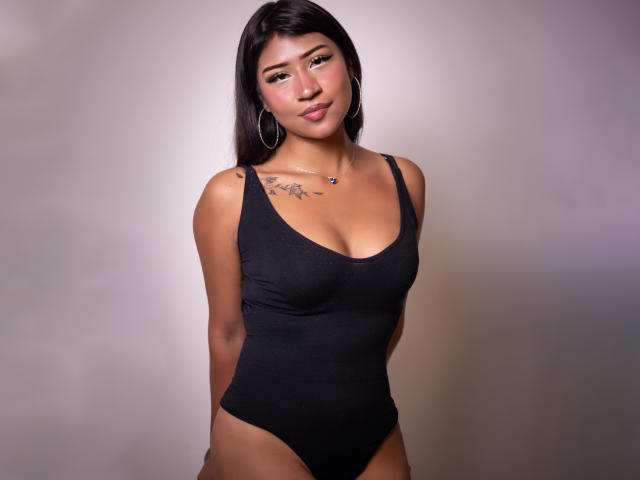BonnieBodn with medium brown skin, black straight hair, wearing a black one-piece, showing her tits, arms behind back, small tattoo on right shoulder, hoop earrings, subtle smile.