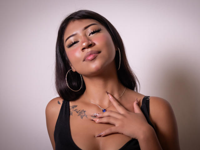 BonnieBodn a la peau mate, des cheveux noirs et lisses, des boucles d'oreilles cerceaux et un débardeur noir. Elle touche sa poitrine, montrant un tatouage et un collier avec un bijou bleu..