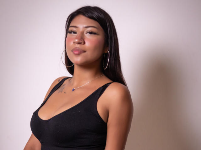 BonnieBodn with medium tits, wearing a black tank top, hoop earrings, and a necklace, has long dark hair, light brown skin, and a slight pout, against a plain background.