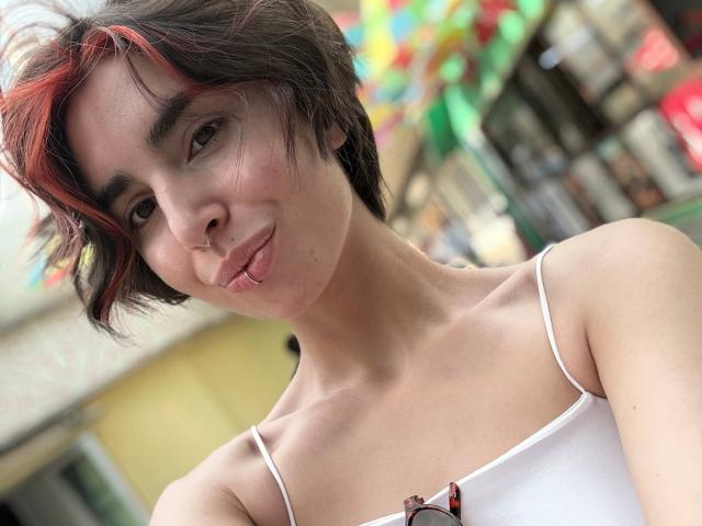 TameikaMacclellan with short brown hair, red highlights, cute nose piercing, white spaghetti strap top, slight pout, and eyeglasses hanging from top. Soft, casual, and slightly sexy.