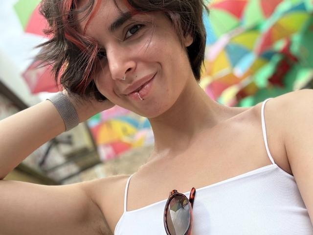 TameikaMacclellan with short brown hair, red highlights, nose ring, lip piercing, white tank top, sunglasses on chest, smiling, gray wristband, armpit visible, colorful background.