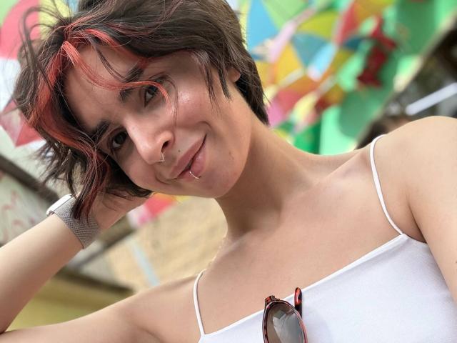TameikaMacclellan with short, dark brown hair with red highlights, white tank top, nose ring, and tongue piercing, smiling at the camera with sunglasses on chest.