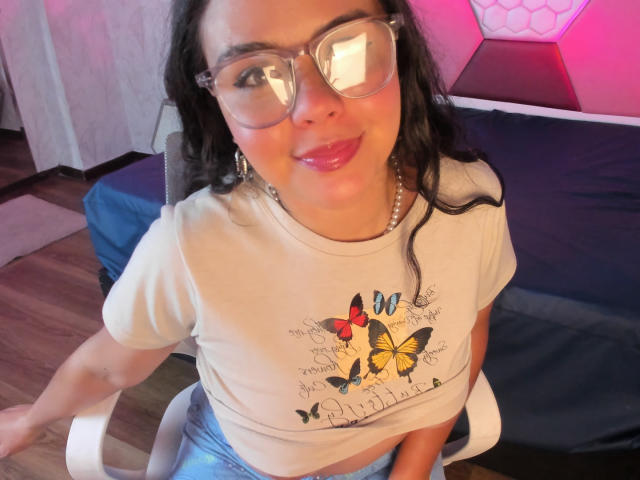 ArielleWilde with glasses, smiling, wearing a white tee with butterfly design, and blue jeans, sitting on a chair, showing her shiny pink lips, and her small tits.