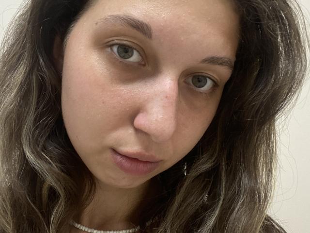 Sanella's face with light brown wavy hair, green eyes, and slightly parted pink lips. She has a small nose and a subtle freckle on her left cheek.