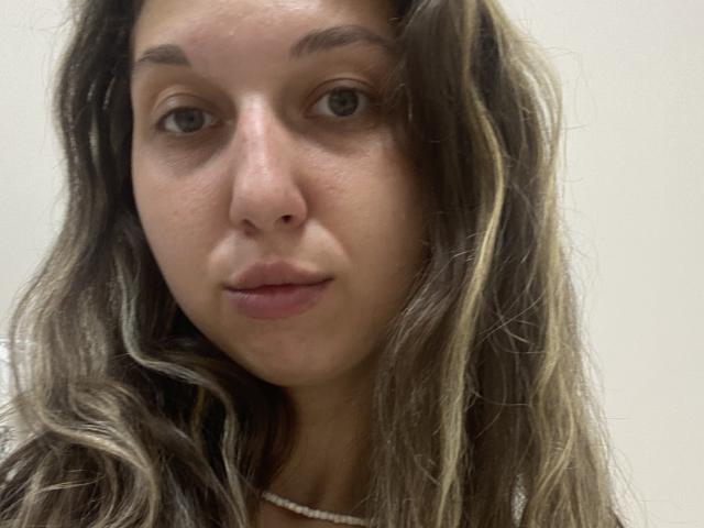 Sanella has long, wavy brown hair with blonde highlights, fair skin, and light green eyes. She's wearing a white necklace and has a neutral expression.