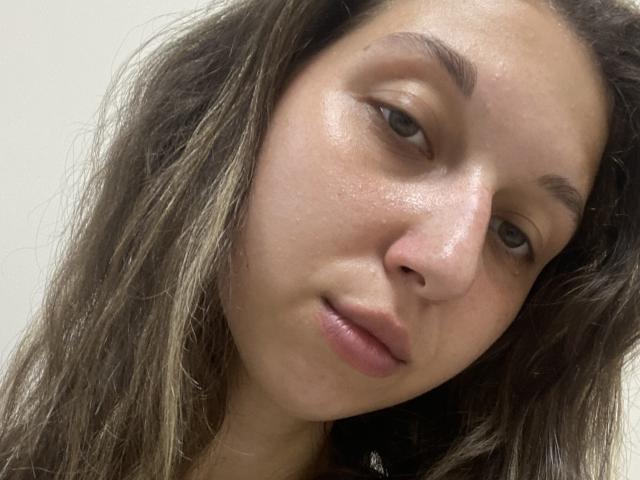 Sanella's face with slightly shiny skin, brown eyes, and parted lips. She has wavy brown hair, small nose, and light brown eyebrows. Her expression is relaxed and neutral.