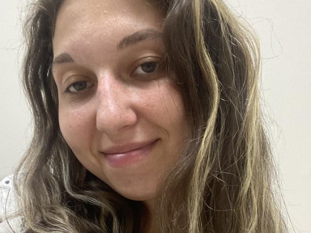 Sanella's face, showing a slight smile with wavy brown and blonde hair, light skin, and subtle freckles. She has a natural, relaxed look with relaxed eyebrows.