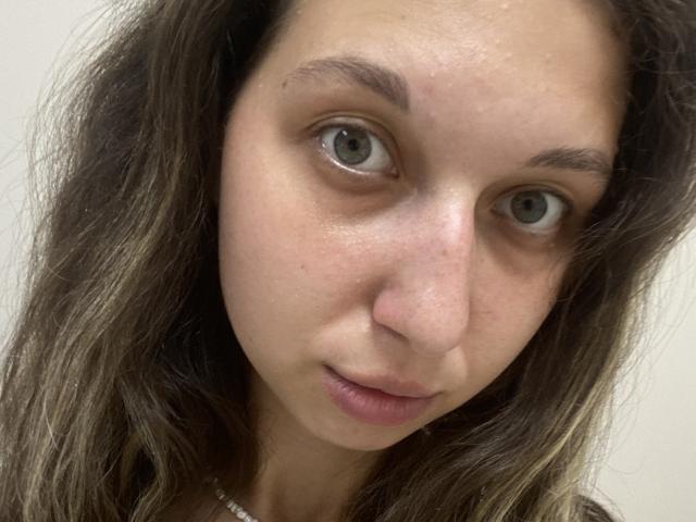 Sanella has a neutral expression, gray eyes, and slightly parted lips. She has wavy brown hair, a fair skin tone, and a small nose. Her eyebrows are slightly raised.